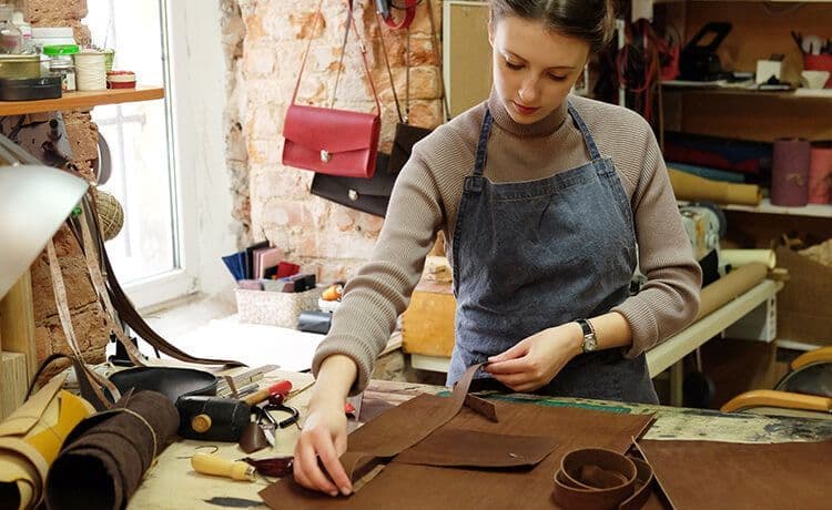 Junge Sattlerin steht in einer Werkstatt und schneidet Lederteile für eine Tasche.
