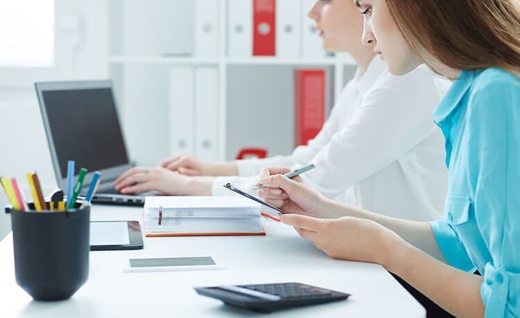 Zwei junge Frauen sitzen an einem Schreibtisch mit Laptop, Stiften, Taschenrechner und Tablet.