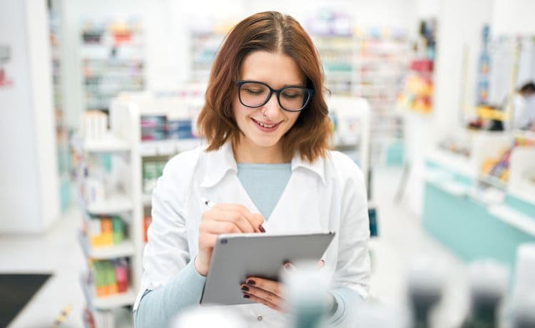 Junge PTA mit Tablet hin der Hand arbeitet in einer Apotheke.