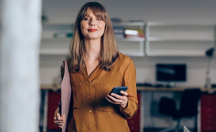 Junge Social Medie Managerin hält ein Smartphone und Unterlagen in den Händen und lächelt in die Kamera.