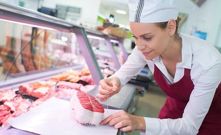 Junge Frau mit Schürze verpackt an der Fleischtheke ein Stück Fleisch.