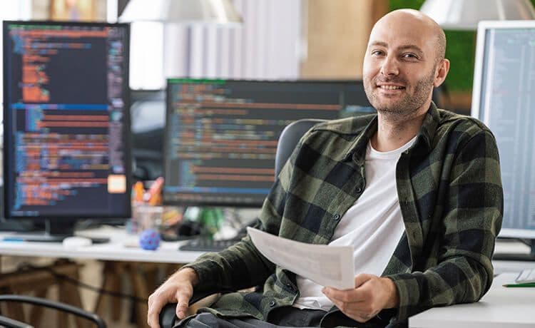 Programmierer sitzt vor einem Computer mit Code und lächelt in die Kamera.