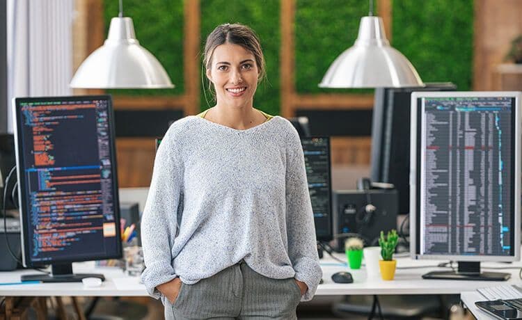 Junge Programmiererin steht in einem Büro vor Computern mit Code und schaut in die Kamera.