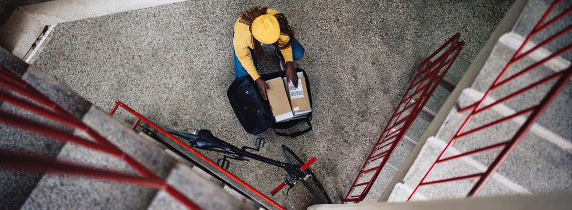 Postbotin sortiert Pakete im Rucksack und kniet dabei im Treppenhaus