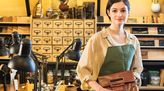 Junge Sattlerin in Ausbildung steht in einer Werkstatt, hält eine Ledertasche in der Hand und lächelt in die Kamera.