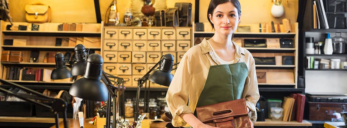 Junge Sattlerin in Ausbildung steht in einer Werkstatt, hält eine Ledertasche in der Hand und lächelt in die Kamera.